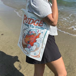 May include: A white canvas tote bag featuring a graphic illustration of an orange fish with blue spots surrounded by bubbles and water waves. The text PUDGE Controls The Weather is printed in orange and blue lettering above the fish illustration.
