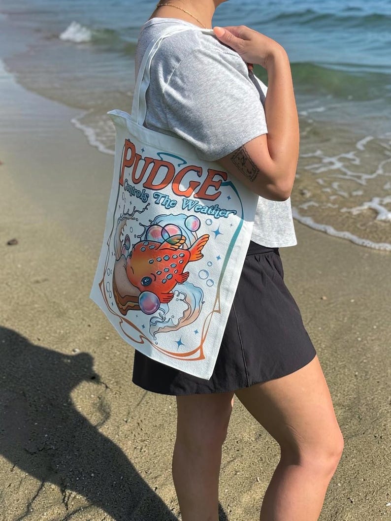 May include: A white canvas tote bag featuring a graphic illustration of an orange fish with blue spots surrounded by bubbles and water waves. The text PUDGE Controls The Weather is printed in orange and blue lettering above the fish illustration.