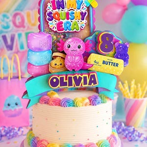 May include: A white frosted cake with rainbow swirl borders and a colourful cake topper featuring the text In My Squishy Era, a pink axolotl, a butter stick, a number 8, and a banner with the name Olivia in purple letters on a teal background.