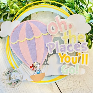 Peut inclure: Une décoration de gâteau en papier superposé représentant une montgolfière violette et rose pastel avec un petit personnage de dessin animé dans la nacelle. Le texte "Oh, the Places You'll Go!" est écrit en lettres multicolores sur un cadre circulaire bleu clair et jaune avec des nuages en papier blanc.