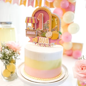 May include: A pink and yellow ombre cake with a white frosting layer. The cake is decorated with a pink and yellow lemonade stand topper with the number 1 and the name Ava.
