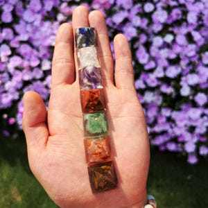 May include: A vertical stack of seven small, square-based pyramid ornaments made of various polished gemstones. The stones display a spectrum of colors including deep blue, clear white, purple, bright red, light green, orange, and dark brown.