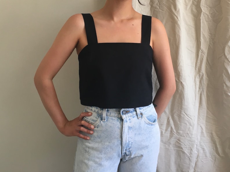 May include: A black cropped tank top with wide straps. The top is made of a smooth, lightweight fabric. The model is wearing the top with light blue denim jeans.