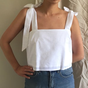 May include: A white sleeveless top with a square neckline and tie straps. The top is cropped and has a relaxed fit.