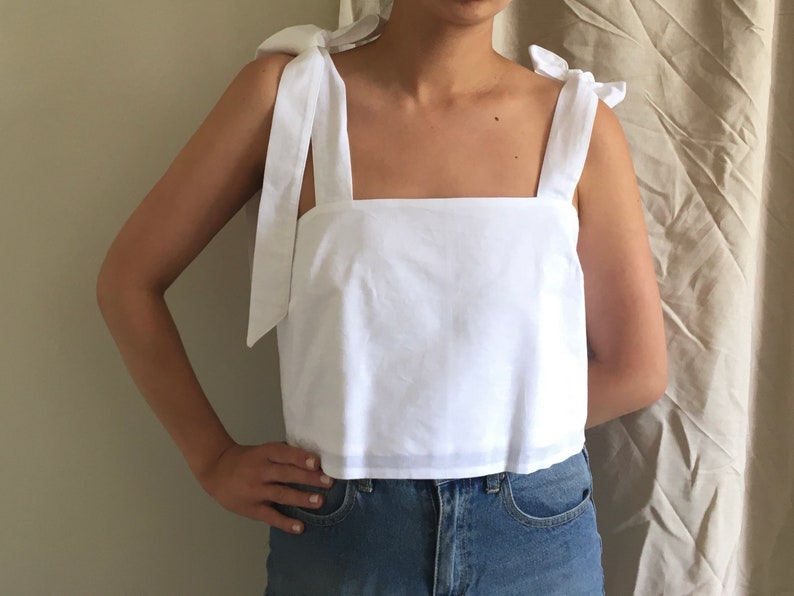 May include: A white sleeveless top with a square neckline and tie straps. The top is cropped and has a relaxed fit.