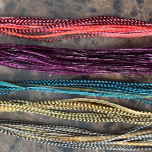 Puede incluir: Cinco paquetes de plumas de pelo coloridas en tonos rosa, morado, turquesa, dorado y gris. Cada paquete está atado con una pequeña banda. Las plumas tienen un patrón moteado y están dispuestas horizontalmente.
