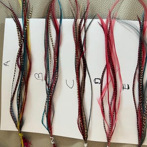 以下が含まれることがあります： 赤、黒、青、黄色など、さまざまな色の羽のヘアエクステンションが白い紙に展示されています。 羽は縦に並べられ、手書きの文字でグループ分けされています。