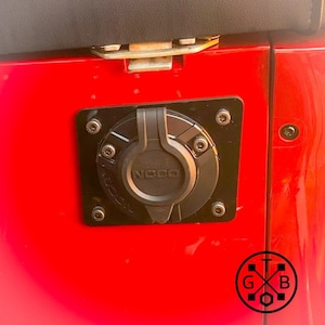 Peut inclure: Un port de charge NOCO noir install&eacute; sur un panneau de v&eacute;hicule rouge. Le port circulaire est dot&eacute; d'un couvercle &agrave; charni&egrave;re et est fix&eacute; par plusieurs vis. Le logo NOCO est visible sur le couvercle. Une plaque carr&eacute;e noire entoure le port.