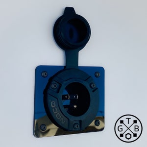 Peut inclure: Une prise d'alimentation NOCO noire avec un capuchon de protection, mont&eacute;e sur une plaque carr&eacute;e bleue r&eacute;fl&eacute;chissante. L'entr&eacute;e a un design circulaire avec des broches visibles et est fix&eacute;e avec des vis. Le fond est une surface blanche unie.