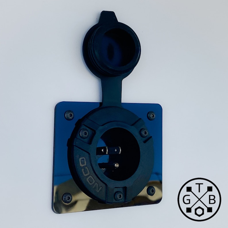 Peut inclure: Une prise d'alimentation NOCO noire avec un capuchon de protection, mont&eacute;e sur une plaque carr&eacute;e bleue r&eacute;fl&eacute;chissante. L'entr&eacute;e a un design circulaire avec des broches visibles et est fix&eacute;e avec des vis. Le fond est une surface blanche unie.