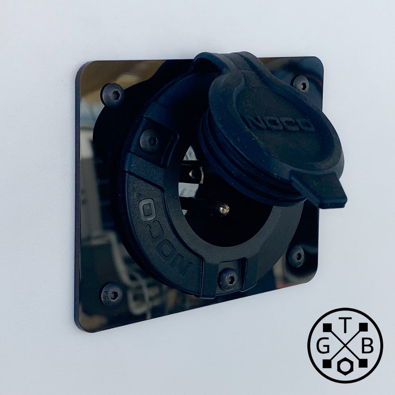 Peut inclure: Une prise de courant noire NOCO mont&eacute;e sur une plaque rectangulaire noire avec quatre vis. La prise a un couvercle de protection et est plac&eacute;e sur un fond blanc. Le logo NOCO est visible sur le couvercle et la lunette de la prise.