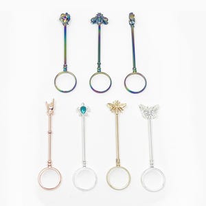 May include: Collection of seven metal finger rings with decorative tops. The rings feature various designs, including a rose, a bee, a skull, a devil horn, a jeweled insect, a bee, and a butterfly. The rings are in various colors, including iridescent, gold, and silver.