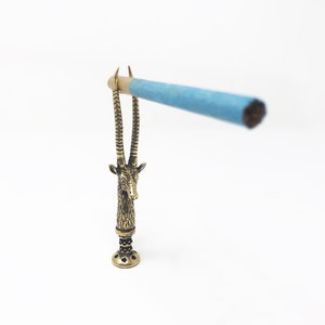 May include: A gold-colored metal incense holder shaped like an antelope head with a blue incense stick resting on the horns.