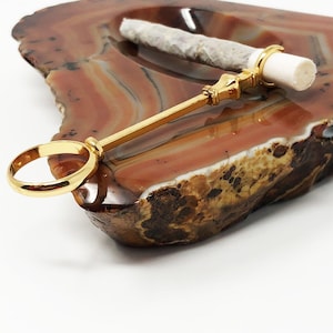 May include: A gold metal smoking tool with a ring on one end and a decorative handle on the other. The tool is resting on a brown and white agate ashtray.