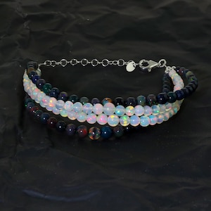 May include: A multi-strand bracelet with a silver chain clasp. The bracelet features three rows of beads: a row of white opal beads, a row of dark blue beads, and a row of iridescent black beads.