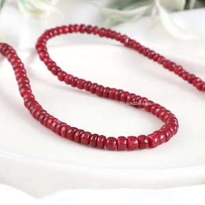 May include: A strand of small, circular, deep red ruby beads arranged in a curved line. The beads have a smooth, polished texture and a consistent size, featuring the text Minedbeads printed in small white letters near the centre of the strand.