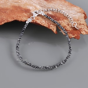 Op de afbeelding: Een verfijnde armband met een doorlopende streng van kleine, donkergrijze ruwe diamantkralen. De armband is voorzien van een zilverkleurige metalen ketting met een karabijnsluiting en een verstelbaar verlengstukje met een klein metalen plaatje.