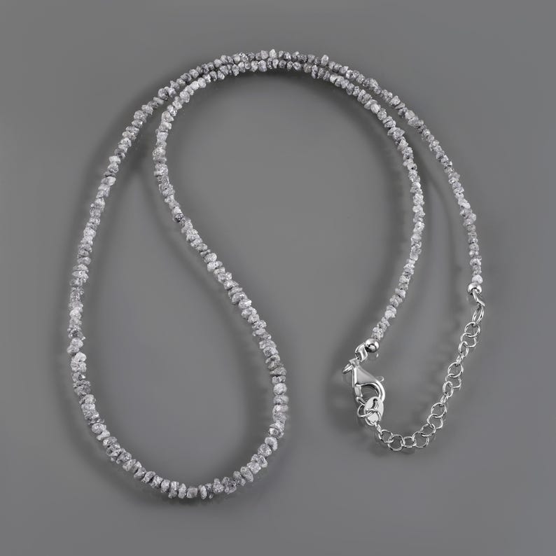 May include: A silver necklace made of small, rough, grey diamonds. The necklace has a lobster clasp closure.