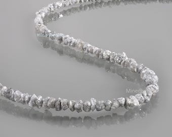 Könnte beinhalten: Ein Strang aus rohen, ungeschliffenen grauen Diamanten, die in einer geschwungenen Linie angeordnet sind. Die kleinen, unregelmäßigen und texturierten Steine haben ein natürliches, facettiertes Aussehen mit metallischem Glanz. Der Text Minedbeads ist sichtbar.