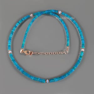 May include: A necklace composed of small, circular blue and green beads with occasional white accent beads. The necklace features a rose gold-toned metal lobster claw clasp and an adjustable extender chain. The text Minedbeads is visible.