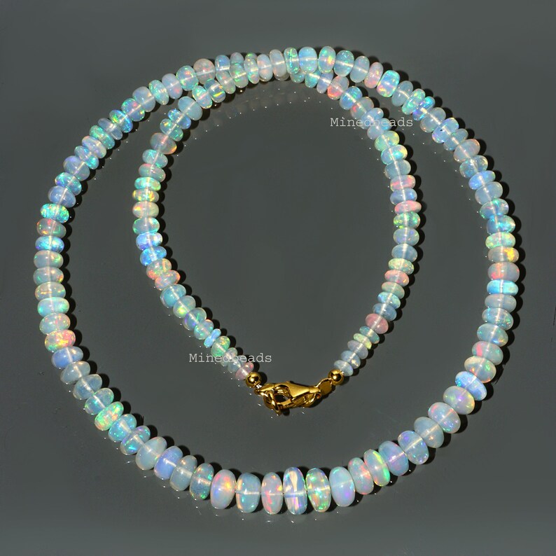 K&ouml;nnte beinhalten: Eine Halskette bestehend aus zahlreichen kleinen, runden, durchscheinenden Opalperlen, die irisierende Blitze in Blau, Gr&uuml;n und Rosa zeigen. Die Kette wird mit einem polierten, goldfarbenen Karabinerverschluss aus Metall mit dem Text Minedbeads geschlossen.