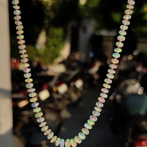 Pode incluir: Um colar feito de contas de opala iridescente. As contas s&atilde;o de forma oval e t&ecirc;m um tom rosa claro. O colar est&aacute; pendurado na frente de um fundo desfocado.