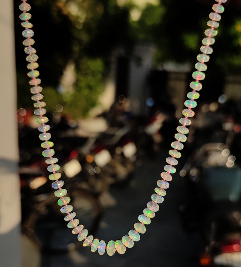 Pode incluir: Um colar feito de contas de opala iridescente. As contas s&atilde;o de forma oval e t&ecirc;m um tom rosa claro. O colar est&aacute; pendurado na frente de um fundo desfocado.