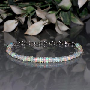 May include: A delicate bracelet featuring a single strand of small, round, translucent opal beads with iridescent flashes of color. The bracelet includes a silver-toned metal chain with a lobster claw clasp and the text Minedbeads. visible twice.