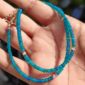 May include: A beaded necklace featuring small, round, vibrant blue-green gemstone beads strung together. The necklace includes metallic gold-toned spacers and small, pale, iridescent accent beads. A gold-toned metal chain extender with a clasp is attached.