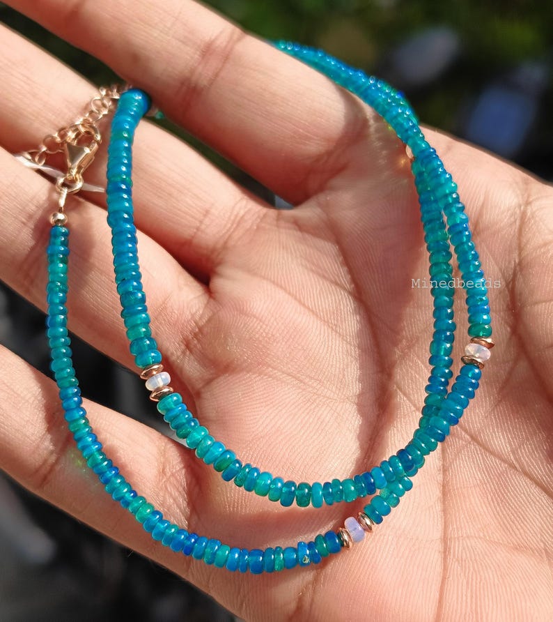 May include: A beaded necklace featuring small, round, vibrant blue-green gemstone beads strung together. The necklace includes metallic gold-toned spacers and small, pale, iridescent accent beads. A gold-toned metal chain extender with a clasp is attached.