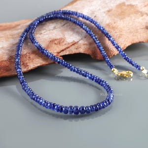 May include: A necklace composed of small, round, deep blue sapphire beads arranged in a single strand. The necklace features a polished gold-toned metal lobster claw clasp and jump ring closure, providing a secure connection for the gemstone strand.