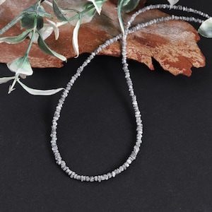 May include: A silver necklace made of small, rough, grey diamonds. The necklace is on a brown, textured surface.