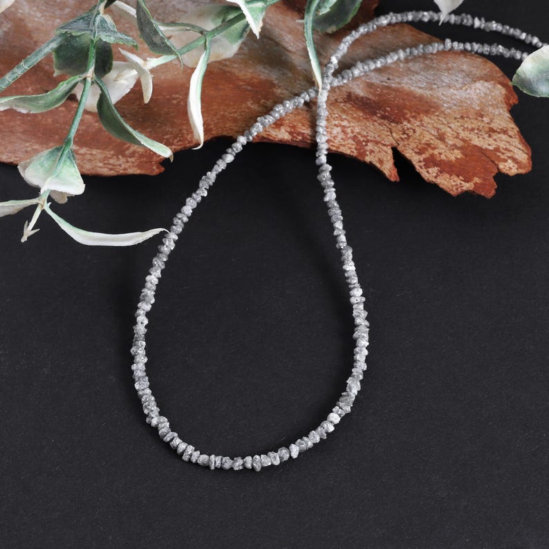 May include: A silver necklace made of small, rough, grey diamonds. The necklace is on a brown, textured surface.