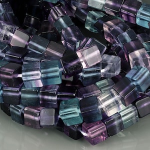 May include: A collection of cube-shaped beads in shades of purple, teal, and clear. The beads are strung together, showcasing their multifaceted design and translucent quality. The beads are likely for jewelry making.
