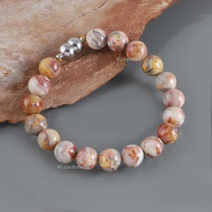 Puede incluir: Una pulsera de cuentas con piedras de ágata redondas y multicolores con patrones en espiral en tonos crema, bronceado, marrón y naranja. La pulsera se asegura con un cierre magnético plateado pulido e incluye el texto Minedbeads.