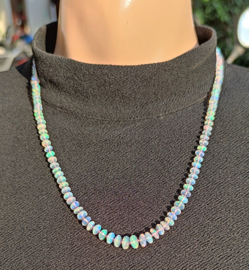 Pode incluir: Um colar feito de contas de opala ovaladas iridescentes. O colar est&aacute; sobre um fundo preto.