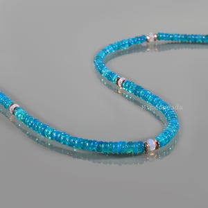 May include: A strand of small, circular, vibrant blue beads arranged in a curved line. The necklace features occasional accents of translucent, milky white beads separated by thin, metallic copper-colored spacers. The text Minedbeads is visible.