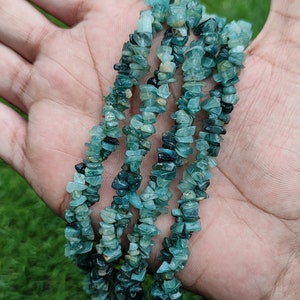 May include: A strand of blue green gemstone chips, also known as teal, arranged in a wavy pattern on a hand. The chips are irregular in shape and size.
