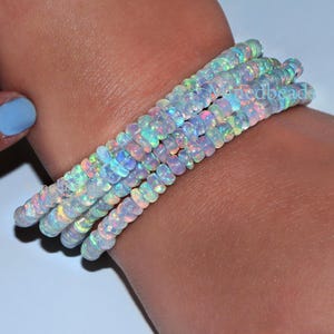 Op de afbeelding: Een armband met meerdere strengen, bestaande uit kleine, ronde, doorschijnende opaal kralen. Elke kraal vertoont een kleurenspel met flitsen van iriserend groen, blauw en roze licht. De kralen zijn geregen in een doorlopend cirkelvormig patroon.