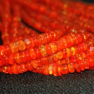 May include: A strand of small, round, vibrant orange fire opal beads with subtle iridescent flashes of yellow and green. The beads are strung together in a continuous line, and the text Minedbeads is visible on the right side of the image.