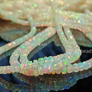 May include: A strand of faceted, translucent white opal beads featuring a vibrant play-of-color with flashes of green, orange, and blue. The beads are circular and disc-shaped, arranged in a loose coil. The text Minedbeads is visible.