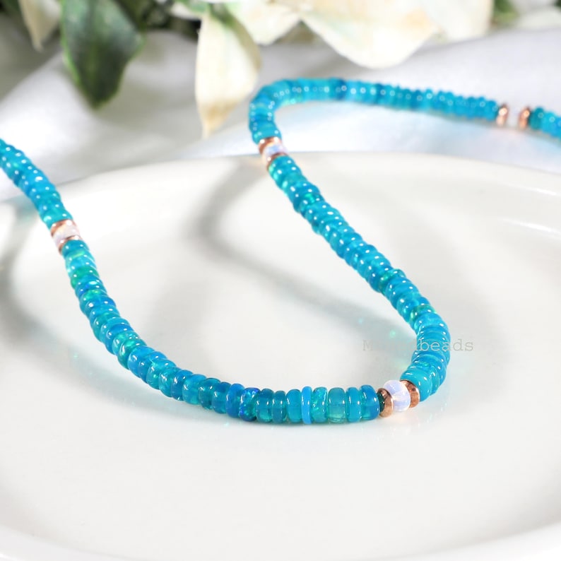 May include: A necklace featuring small, vibrant turquoise blue disc-shaped beads arranged in a continuous strand. The design is punctuated at regular intervals by smooth, round, iridescent white beads flanked by thin, metallic copper-colored spacers.