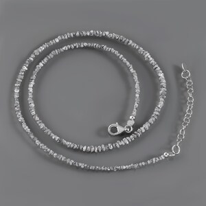May include: A silver necklace made of small, rough, gray diamonds. The necklace is on a gray background.