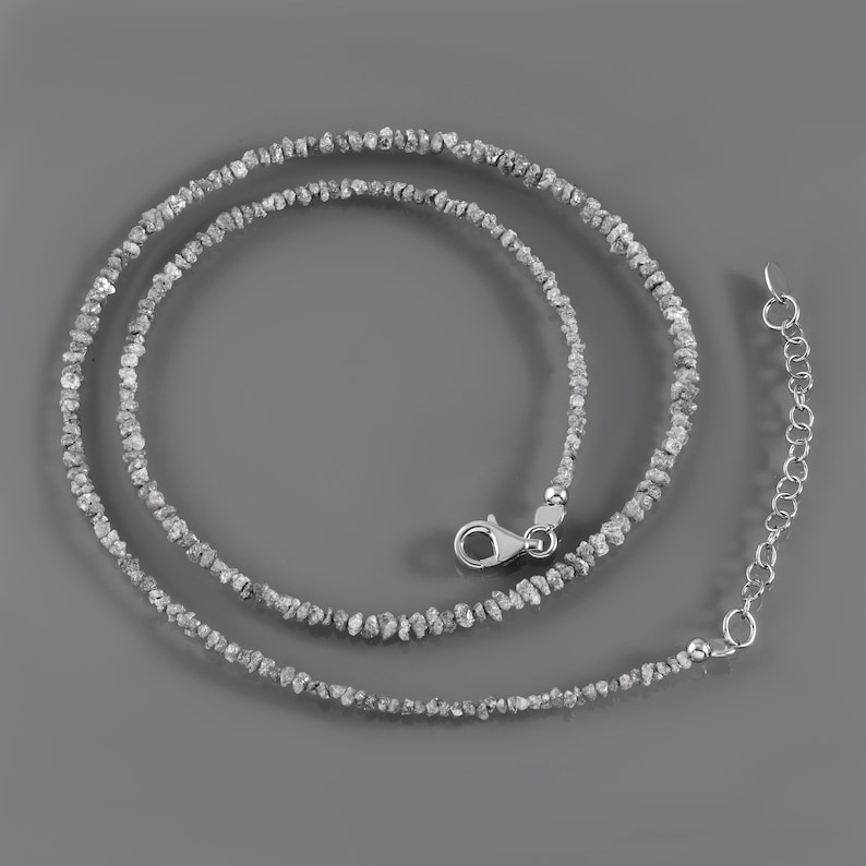 May include: A silver necklace made of small, rough, gray diamonds. The necklace is on a gray background.