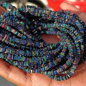 Puede incluir: Una hebra de cuentas de ópalo iridiscente, cada cuenta es de forma rectangular con un color azul oscuro, verde y morado.