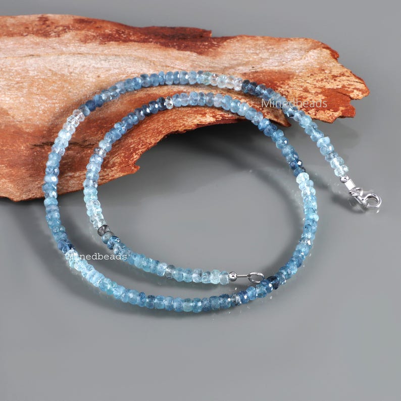 May include: A necklace composed of small, faceted, round blue gemstone beads that transition in color from light sky blue to deep navy blue. The strand features a silver-toned metal lobster claw clasp and small silver-toned metal spacer beads.