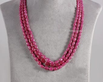 May include: A multi-strand necklace featuring three layers of smooth, oval-shaped, vibrant pink beads. The beads are uniform in size and polished to a glossy finish. The text Minedbeads is visible on the right side of the necklace strands.