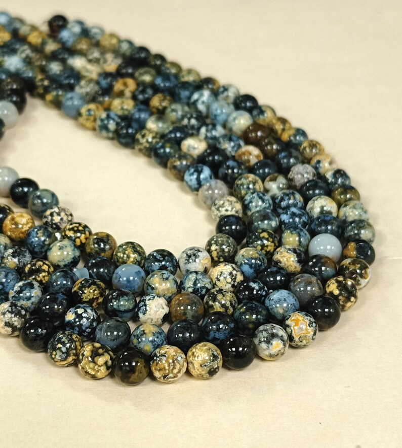 May include: A strand of blue, brown, and white speckled round beads. The beads are made of natural stone and have a mottled appearance.