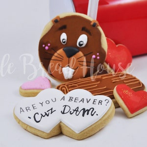 May include: A collection of decorated sugar cookies, including a beaver-shaped cookie, heart-shaped cookies, and log-shaped cookies. The cookies are arranged near a red mailbox. One cookie reads "Are you a beaver? 'Cuz DAM."