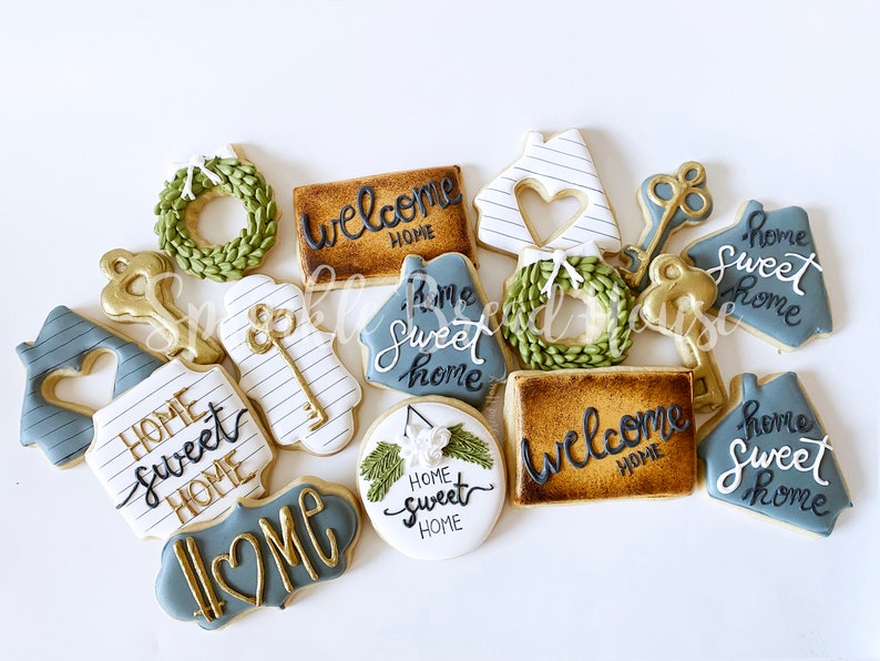 Puede incluir: Surtido de galletas decoradas con dise&ntilde;os tem&aacute;ticos de hogar. Las galletas incluyen formas de casa, llaves, coronas y letreros con frases como "Welcome Home" y "Home Sweet Home". Los colores incluyen dorado, blanco y tonos de azul y verde.